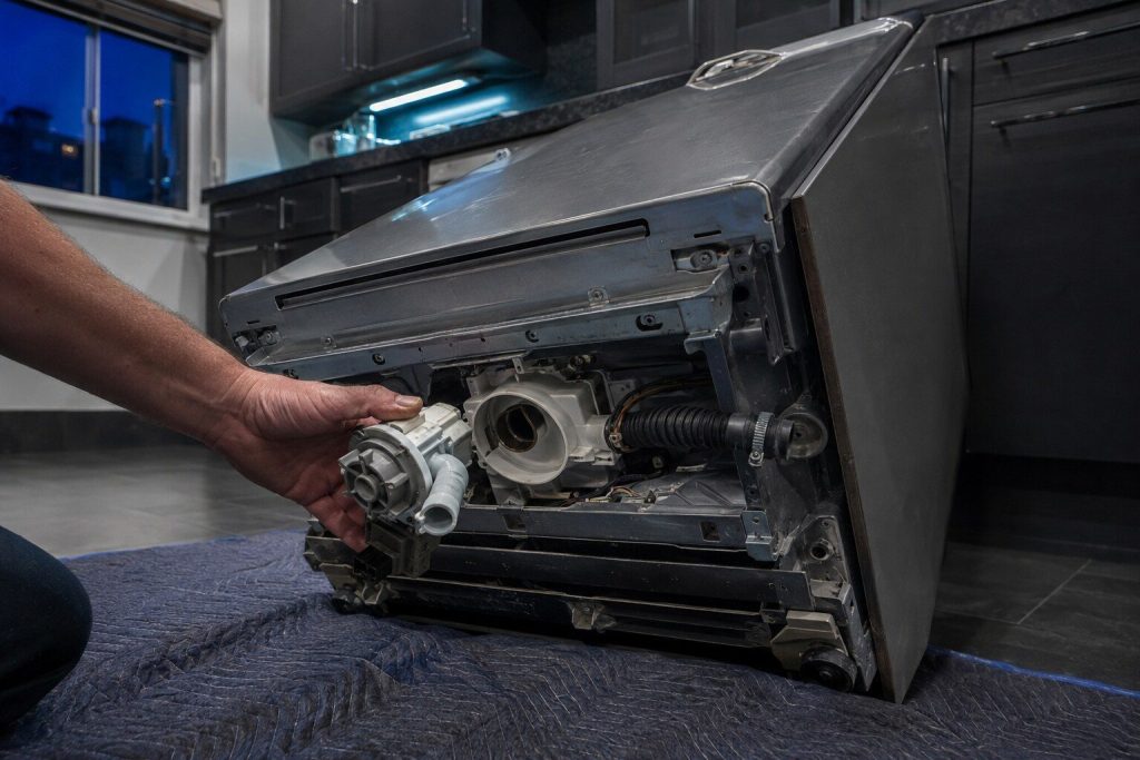 A dishwasher lying tipped back on a blanket with its lower access panel off and the drain pump housing exposed on the underside, the pump held in one hand.