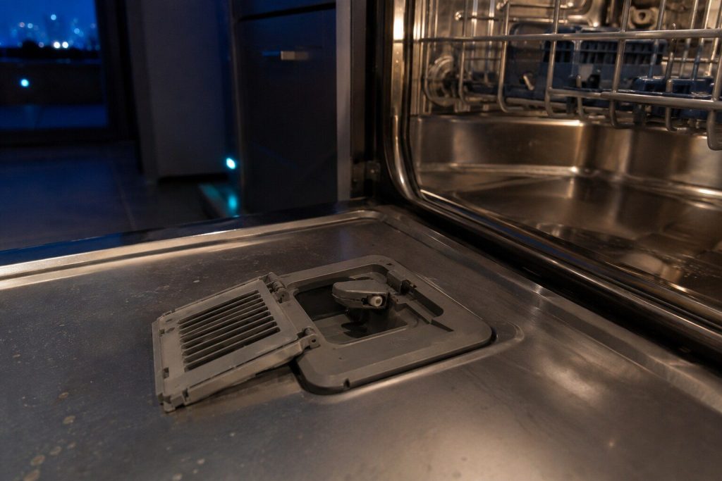 The drying vent in the inner face of an open dishwasher door with its cover unclipped and lifted aside, the small flap and its actuator visible in the recess.