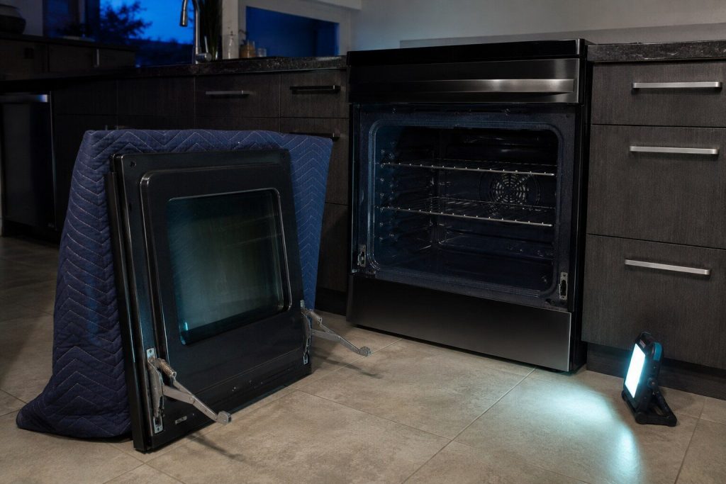 An oven door lifted off the range and stood upright against a blanket on the floor, both hinge arms locked open, the empty hinge slots in the frame behind it.