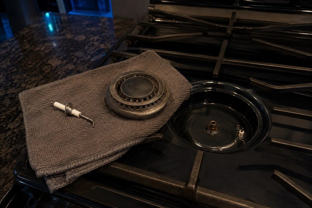 A spark igniter and a burner head lying apart on a folded towel on a cooktop, the empty burner base and its orifice visible beside them.