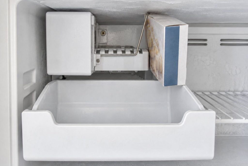 The inside of a freezer compartment with the ice bin pulled out and completely empty, the white ice maker module above it with its wire shut-off arm raised in the up position and a box of frozen food pressed against it.